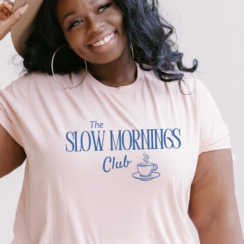 K&ouml;nnte beinhalten: Hellrosa T-Shirt mit dem Text "The Slow Mornings Club" in Dunkelblau. Darunter befindet sich eine kleine Kaffeetassen-Grafik. Das T-Shirt hat einen Rundhalsausschnitt.