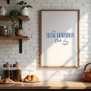 K&ouml;nnte beinhalten: Ein gerahmter Druck mit dem blauen Text "The Slow Mornings Club" und einem Kaffeetassen-Symbol. Der Druck h&auml;ngt an einer wei&szlig;en Backsteinmauer &uuml;ber einer Holztheke mit Croissants, Gl&auml;sern und Regalen mit Pflanzen.