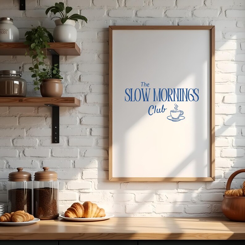 K&ouml;nnte beinhalten: Ein gerahmter Druck mit dem blauen Text "The Slow Mornings Club" und einem Kaffeetassen-Symbol. Der Druck h&auml;ngt an einer wei&szlig;en Backsteinmauer &uuml;ber einer Holztheke mit Croissants, Gl&auml;sern und Regalen mit Pflanzen.