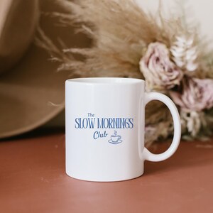 K&ouml;nnte beinhalten: Wei&szlig;e Keramik-Tasse mit dem blauen Schriftzug "The Slow Mornings Club". Darunter eine Illustration einer Tasse mit Untertasse. Die Tasse hat einen gebogenen Henkel. Der Hintergrund ist unscharf mit Trockenblumen.