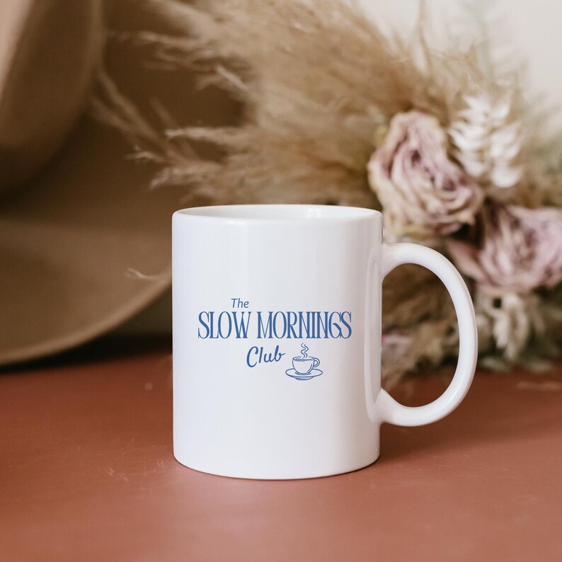 K&ouml;nnte beinhalten: Wei&szlig;e Keramik-Tasse mit dem blauen Schriftzug "The Slow Mornings Club". Darunter eine Illustration einer Tasse mit Untertasse. Die Tasse hat einen gebogenen Henkel. Der Hintergrund ist unscharf mit Trockenblumen.