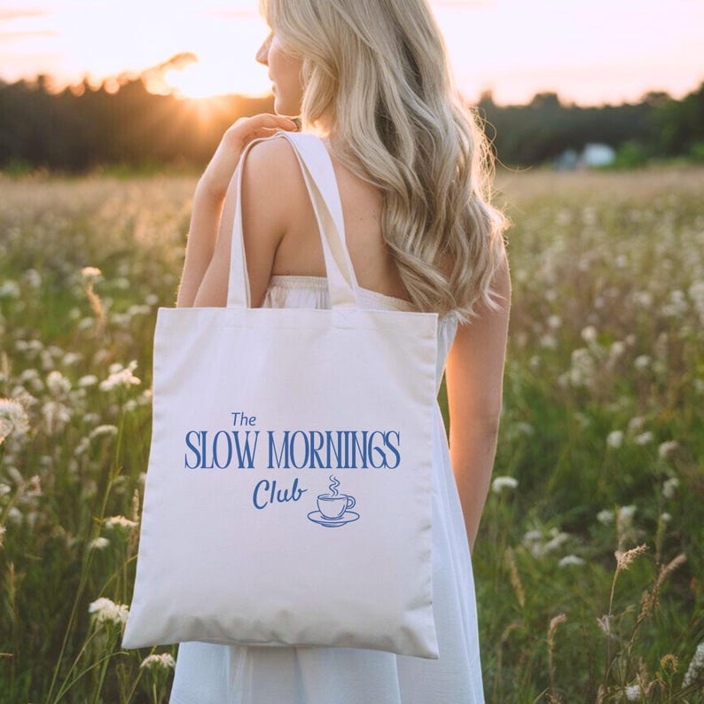 K&ouml;nnte beinhalten: Eine wei&szlig;e Tragetasche mit dem blauen Text "The Slow Mornings Club" und einer Kaffeetassen-Illustration. Die Tasche wird von einer Person in einem wei&szlig;en Kleid getragen, mit einem Feld und Sonnenuntergang im Hintergrund.