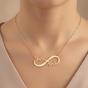 Può includere: Una collana color oro con un ciondolo a forma di simbolo dell'infinito. Il ciondolo ha due nomi, "Jerber" e "Honbun", scritti in corsivo. La collana è indossata intorno al collo.