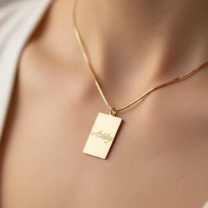 May include: A gold-colored rectangular pendant necklace with the name "Ashley" engraved in a cursive font. The pendant hangs from a delicate gold chain, creating a simple and elegant look. The necklace is shown on a person.