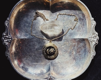 May include: A gold-toned locket necklace featuring a circular pendant with a black inlay depicting a sun and crescent moon. The necklace is accompanied by a clear, pointed quartz crystal, both resting on a silver-toned, four-sectioned metal tray.