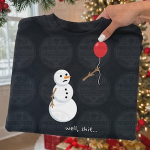 May include: A black crewneck sweatshirt featuring a graphic of a white snowman with a sad expression, three black buttons, and stick arms. One arm holds a red balloon by a string. The text "well, shit..." is printed in white below the snowman.