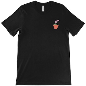 以下が含まれることがあります： ピンクの花が付いた鉢植えの蘭の小さな刺繍デザインが特徴の黒いTシャツ。テラコッタ色の鉢は、暗い生地と対照的です。植物愛好家のためのシンプルでミニマリストなデザイン。