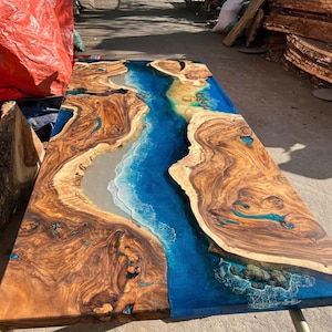 May include: A large, rectangular wooden table with a river-like design. The table features a rich, brown wood grain with a blue epoxy resin river running through the center. The resin mimics ocean waves and a sandy beach.