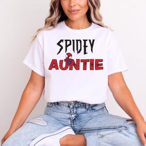 K&ouml;nnte beinhalten: Wei&szlig;es T-Shirt mit dem Text "SPIDEY AUNTIE" in Schwarz und Rot, mit einer Spider-Man-Grafik. Das Shirt ist aus einem weichen Material und eignet sich f&uuml;r Freizeitkleidung. Das Design ist mittig auf der Vorderseite des Shirts platziert.