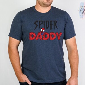 K&ouml;nnte beinhalten: Ein marineblaues T-Shirt mit dem Text "SPIDER DADDY" in Schwarz und Rot. Das Wort "SPIDER" ist in einer stilisierten Schriftart gehalten, und das Wort "DADDY" ist rot mit einem spinnennetzartigen Design. Zwischen den W&ouml;rtern befindet sich eine kleine Spider-Man-Grafik.