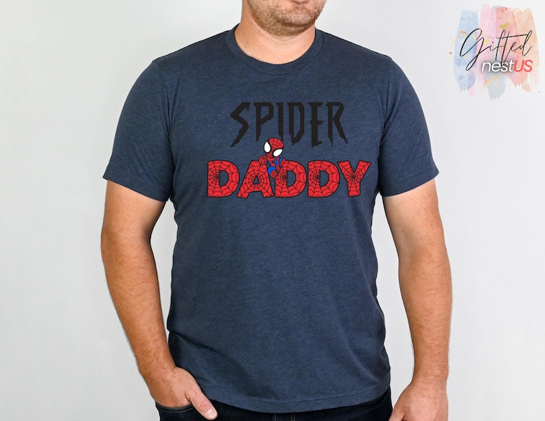 K&ouml;nnte beinhalten: Ein marineblaues T-Shirt mit dem Text "SPIDER DADDY" in Schwarz und Rot. Das Wort "SPIDER" ist in einer stilisierten Schriftart gehalten, und das Wort "DADDY" ist rot mit einem spinnennetzartigen Design. Zwischen den W&ouml;rtern befindet sich eine kleine Spider-Man-Grafik.