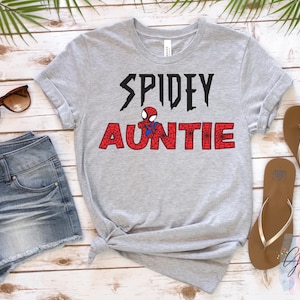 K&ouml;nnte beinhalten: Ein graues T-Shirt mit dem Schriftzug "SPIDEY AUNTIE" in Schwarz und Rot, mit einer Spider-Man-Grafik. Das Bild zeigt Jeansshorts, Sonnenbrille und braune Sandalen auf einer Holzoberfl&auml;che.