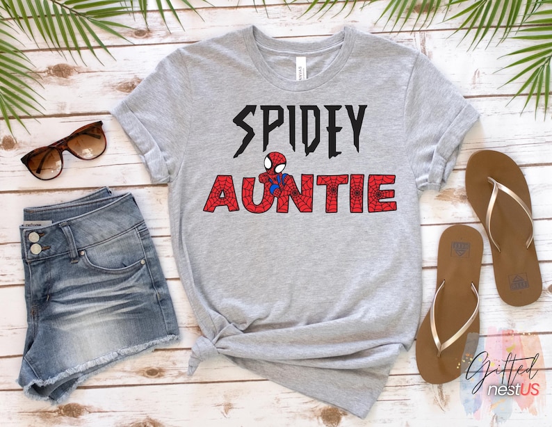 K&ouml;nnte beinhalten: Ein graues T-Shirt mit dem Schriftzug "SPIDEY AUNTIE" in Schwarz und Rot, mit einer Spider-Man-Grafik. Das Bild zeigt Jeansshorts, Sonnenbrille und braune Sandalen auf einer Holzoberfl&auml;che.