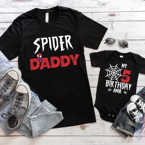 K&ouml;nnte beinhalten: Schwarze Spider-Man-T-Shirts und ein Body. Das Erwachsenen-Shirt tr&auml;gt den Schriftzug "SPIDER DADDY" in wei&szlig;er und roter Schrift. Der Body tr&auml;gt den Schriftzug "MY BIRTHDAY 5 AMIR" mit einem Spinnennetz-Design. Enth&auml;lt Jeans und graue Turnschuhe.