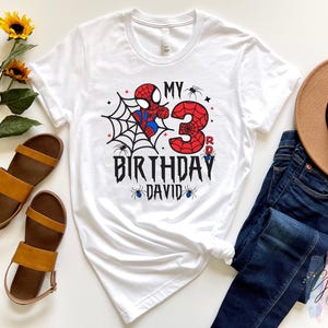 K&ouml;nnte beinhalten: Wei&szlig;es T-Shirt mit einem Spider-Man-Motiv zum dritten Geburtstag. Das Design umfasst ein Spinnennetz, die Zahl 3 und den Text "MY BIRTHDAY DAVID". Braune Sandalen, ein brauner Hut und blaue Jeans sind ebenfalls im Bild zu sehen.