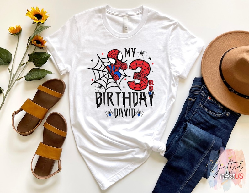 K&ouml;nnte beinhalten: Wei&szlig;es T-Shirt mit einem Spider-Man-Motiv zum dritten Geburtstag. Das Design umfasst ein Spinnennetz, die Zahl 3 und den Text "MY BIRTHDAY DAVID". Braune Sandalen, ein brauner Hut und blaue Jeans sind ebenfalls im Bild zu sehen.