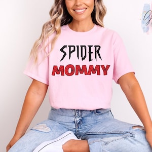 K&ouml;nnte beinhalten: Hellrosa T-Shirt mit dem Aufdruck "SPIDER MOMMY" in Schwarz und Rot. Das T-Shirt hat kurze &Auml;rmel und einen Rundhalsausschnitt. Der Text ist in einer kr&auml;ftigen, stilisierten Schriftart gehalten, wobei das Wort "MOMMY" ein spinnennetzartiges Muster aufweist.