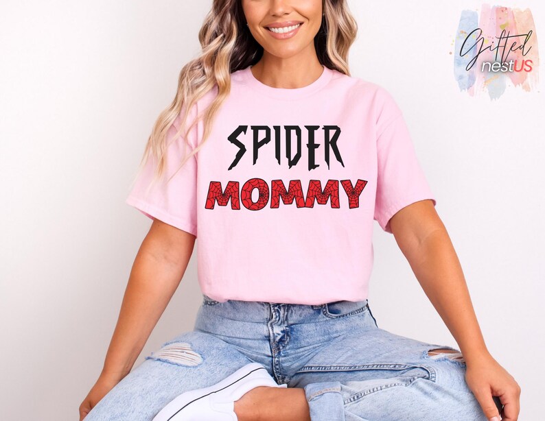 K&ouml;nnte beinhalten: Hellrosa T-Shirt mit dem Aufdruck "SPIDER MOMMY" in Schwarz und Rot. Das T-Shirt hat kurze &Auml;rmel und einen Rundhalsausschnitt. Der Text ist in einer kr&auml;ftigen, stilisierten Schriftart gehalten, wobei das Wort "MOMMY" ein spinnennetzartiges Muster aufweist.