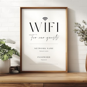 May include: A framed sign with the word "WIFI" in large, bold letters, followed by "For our guests" in a script font. Below are lines for network name and password. The sign is in a wooden frame, set on a wooden surface, with plants and a vase in the background.