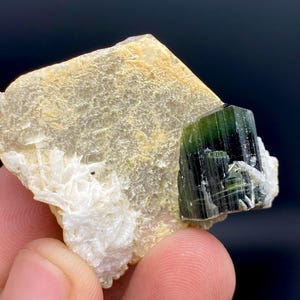 May include: A close-up of a green tourmaline crystal formation on a beige and white mineral base. The tourmaline is a deep green color with visible striations. The base has a rough, textured appearance.