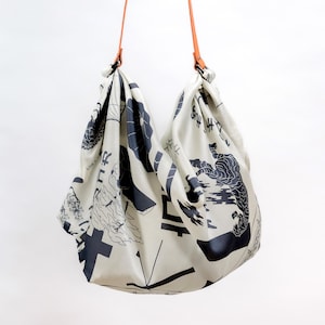 May include: Cream-colored tote bag with a black graphic print featuring Japanese characters and illustrations. The bag has orange leather straps and a unique, soft shape. The design includes a tiger and other abstract shapes.