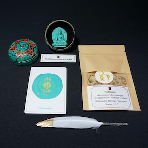 Könnte beinhalten: Eine Ansammlung spiritueller Gegenstände, darunter eine türkisfarbene Buddha-Statue in einer schwarzen Schale, eine dekorative Kugel, eine Karte mit einem türkisfarbenen Design, eine Feder und ein Beutel mit Kräutern. Die Gegenstände sind auf einer dunklen Oberfläche angeordnet.