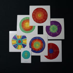 Könnte beinhalten: Eine Sammlung quadratischer Kunstdrucke mit farbenfrohen, kreisförmigen Mandala-Designs. Jeder Druck zeigt eine einzigartige Farbkombination, darunter Rot, Orange, Gelb, Grün, Blau und Lila, mit goldenen Akzenten. Auf schwarzem Hintergrund angeordnet.