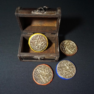 Könnte beinhalten: Eine kleine, offene Holzschatztruhe mit silbernem Verschluss enthält eine große, runde, silberfarbene Münze mit gelbem Rand. Drei zusätzliche Münzen mit Pentagramm-Design und farbigen Rändern (orange, braun und blau) befinden sich in der Nähe.