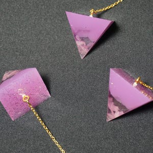 Könnte beinhalten: Drei geometrische Anhänger-Halsketten. Jede Halskette hat einen lila, dreieckigen Anhänger mit Glitzerakzenten und einer goldfarbenen Kette. Die Anhänger haben ein geschichtetes Design mit einem helleren, strukturierten Abschnitt unten.