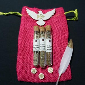 Puede incluir: Una bolsa de yute rosa con tres viales de vidrio llenos de hierbas, cada uno etiquetado en alemán. También hay un recorte de madera en forma de paloma blanca y una pluma blanca con punta dorada. Tres pequeñas fichas redondas blancas con símbolos verdes están en la parte inferior.