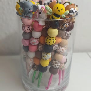 Könnte beinhalten: Ein durchsichtiger Glasbehälter gefüllt mit mehreren Häkelnadeln, die mit dekorativen Silikonperlen in verschiedenen Farben, Mustern und Formen verziert sind, einschließlich Miniaturfiguren von Elefant, Einhorn, Biene und Faultier am Griff.