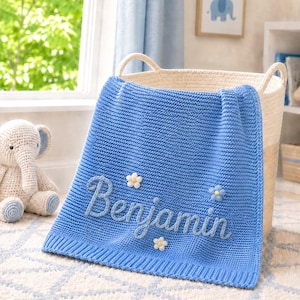Op de afbeelding: Een blauwe gebreide deken met de naam Benjamin geborduurd in cursief schrift, versierd met kleine witte en blauwe bloemapplicaties. De deken heeft een geribbelde rand en hangt over een hoge, ronde, crèmekleurige gevlochten mand.