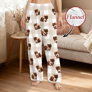 May include: Flannel pajama pants with a beige and white checkered pattern. The pants are decorated with repeating images of a brown and white dog's face. The word "Flannel" is in a circle in the upper right corner.