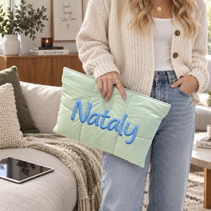 May include: A light green quilted rectangular pouch featuring the name Nataly embroidered in thick, textured blue yarn across the centre. The pouch has a soft, padded fabric construction with visible vertical stitching lines throughout the surface.