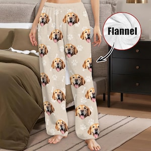 May include: Beige flannel pajama pants featuring a repeating pattern of Golden Retriever faces, white paw prints, and bone shapes. The pants have a relaxed fit and are made of a soft, cozy material.