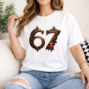 May include: White t-shirt featuring the number 67 in an ornate, vintage-style design. The numbers are dark brown with gold accents and a heart detail on the number 7. The shirt is a casual, everyday wear item.