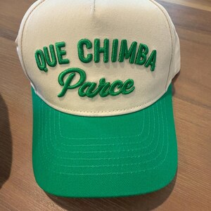 May include: A baseball cap with a beige crown and a green bill. The cap features the embroidered text "QUE CHIMBA Parce" in green. The cap has a green button on top.