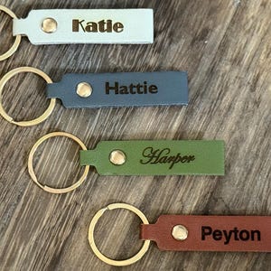 May include: Four personalized leather keychains in various colors: light blue, teal, green, and brown. Each keychain features a gold-colored ring and a name engraved in a contrasting color: Katie, Hattie, Harper, and Peyton.