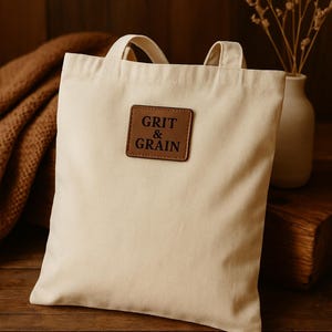 May include: Cream-colored tote bag with short handles and a brown leather patch that reads "GRIT & GRAIN." The bag is sitting on a wooden surface. A vase with dried flowers is in the background.
