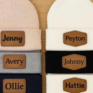 May include: A collection of six personalized beanies in various colors: light pink, white, gray, navy, black, and white. Each beanie features a brown leather patch with a name: Jenny, Peyton, Avery, Johnny, Ollie, and Hattie.