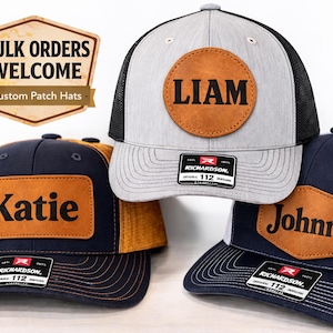 May include: Three personalized trucker hats with leather patches. One hat is gray with a black mesh back and a round patch that says "LIAM". Another is navy blue with a gold mesh back and a rectangular patch that says "Katie". The third is navy blue with a white mesh back and a rectangular patch that says "Johnny".