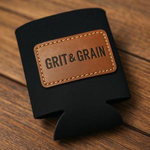 May include: Black neoprene beverage holder with a brown leather patch. The patch is stitched with white thread and features the text "GRIT & GRAIN". The holder is designed to keep drinks cold and is suitable for cans and bottles.