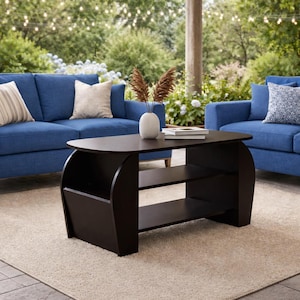 May include: A dark brown coffee table with a distinctive design, featuring a curved top and two shelves. The table is placed on a beige rug in front of two blue sofas with decorative cushions. A white vase with dried plants sits on the table.