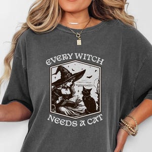 Pode incluir: T-shirt cinzenta escura com o texto "EVERY WITCH NEEDS A CAT" e um gráfico de uma bruxa e um gato preto. A camisa tem um ajuste solto e mangas curtas. A bruxa está usando um chapéu e está sentada ao lado de um gato preto.