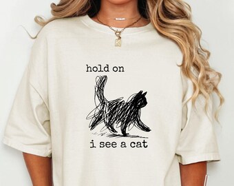 May include: Cream-colored t-shirt featuring a black scribbled cat illustration with the text "hold on i see a cat". The shirt has short sleeves and a relaxed fit.
