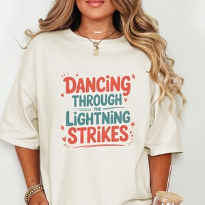 Könnte beinhalten: Cremefarbenes T-Shirt mit dem Text "DANCING THROUGH THE LIGHTNING STRIKES" in roter und türkisfarbener Schrift. Das Shirt hat kurze Ärmel und eine lockere Passform. Die Person hält ein Glas mit einem Strohhalm.