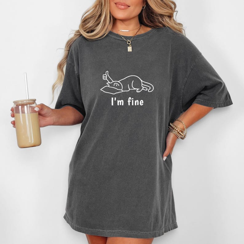May include: Dark grey t-shirt featuring the text "I'm fine" and a white graphic of a cat with a thumbs up. The shirt has a relaxed fit and short sleeves. The model is holding a glass of a light brown beverage.