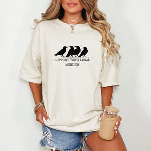 Puede incluir: Camiseta color crema con un gr&aacute;fico negro de cuatro cuervos posados en una l&iacute;nea, con el texto "SUPPORT YOUR LOCAL MURDER" debajo. La camiseta tiene mangas cortas y un corte relajado, adecuada para uso casual.