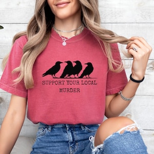 Puede incluir: Camiseta granate con la frase "SUPPORT YOUR LOCAL MURDER" impresa en negro. El dise&ntilde;o presenta cuatro siluetas negras de cuervos. La camiseta es de estilo casual.