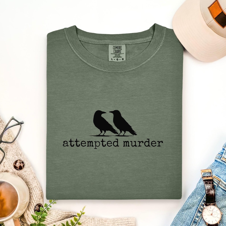 Pu&ograve; includere: Maglietta a maniche lunghe verde salvia con la scritta "attempted murder" sotto una grafica di due corvi neri. La maglietta &egrave; del marchio Comfort Colors, taglia L-G-G.